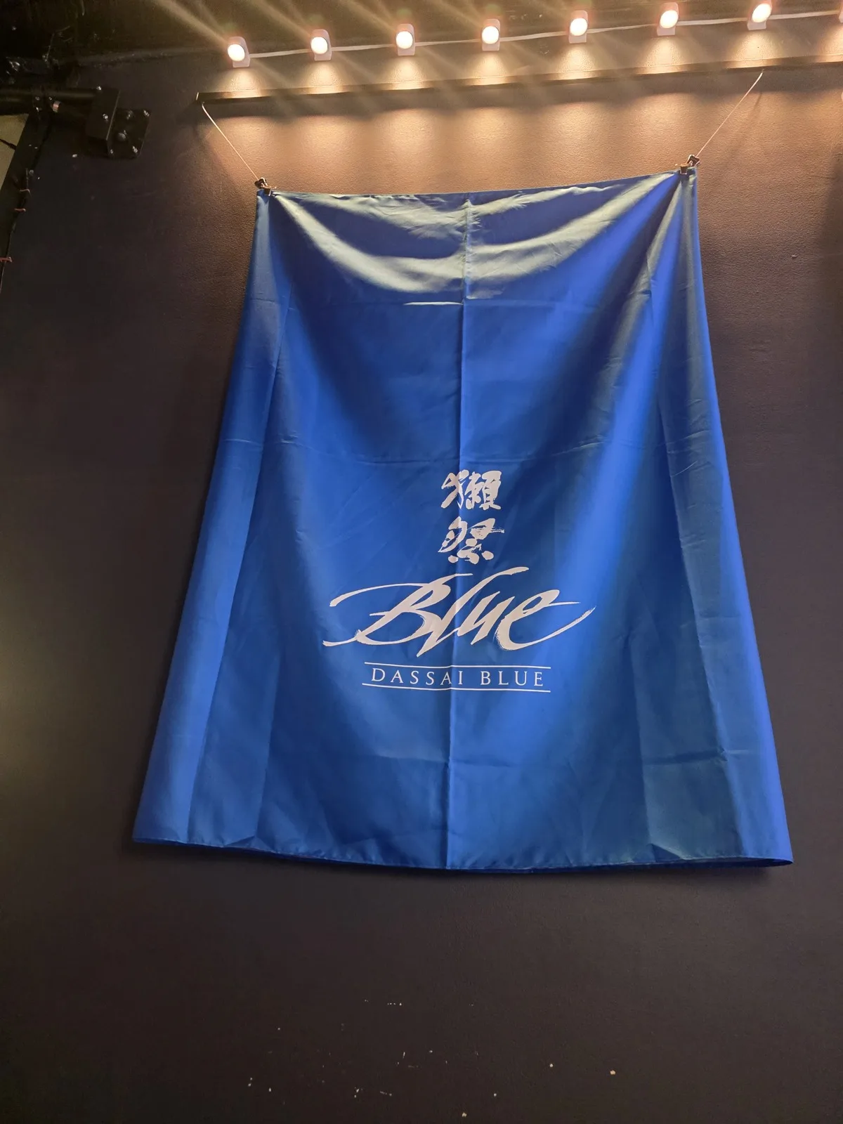 A blue Dassai Blue noren banner hanging on the wall at Izakaya Juraku beneath warm string lights